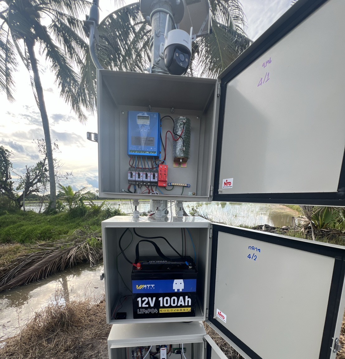 Two open station cabinets in a paddy: charge controller, surge protection and breakers above, and the battery below